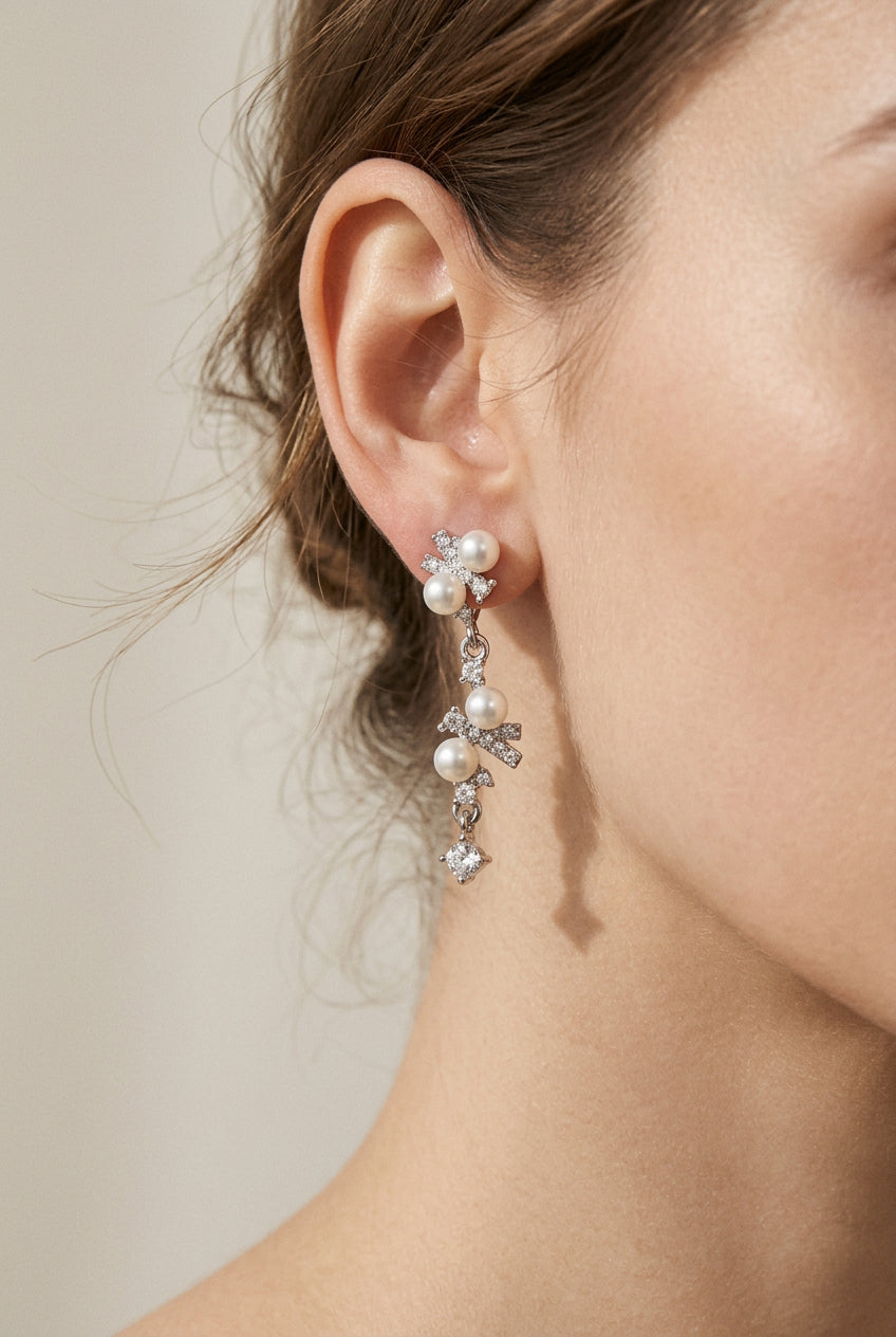 A pair of delicate dangle earrings featuring small white pearls and clear rhinestones on a plain beige background.