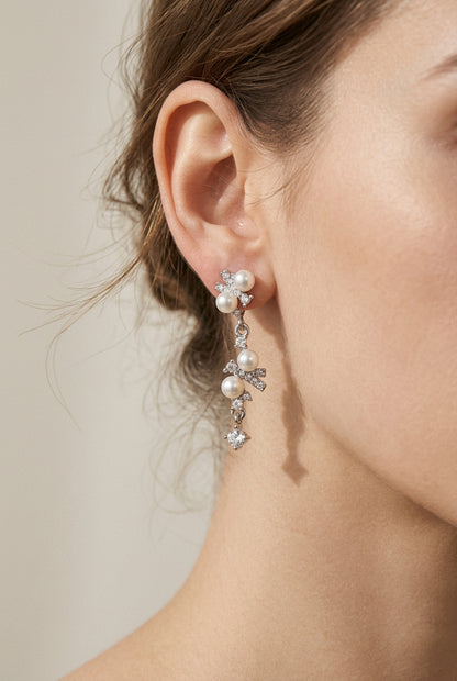 A pair of delicate dangle earrings featuring small white pearls and clear rhinestones on a plain beige background.